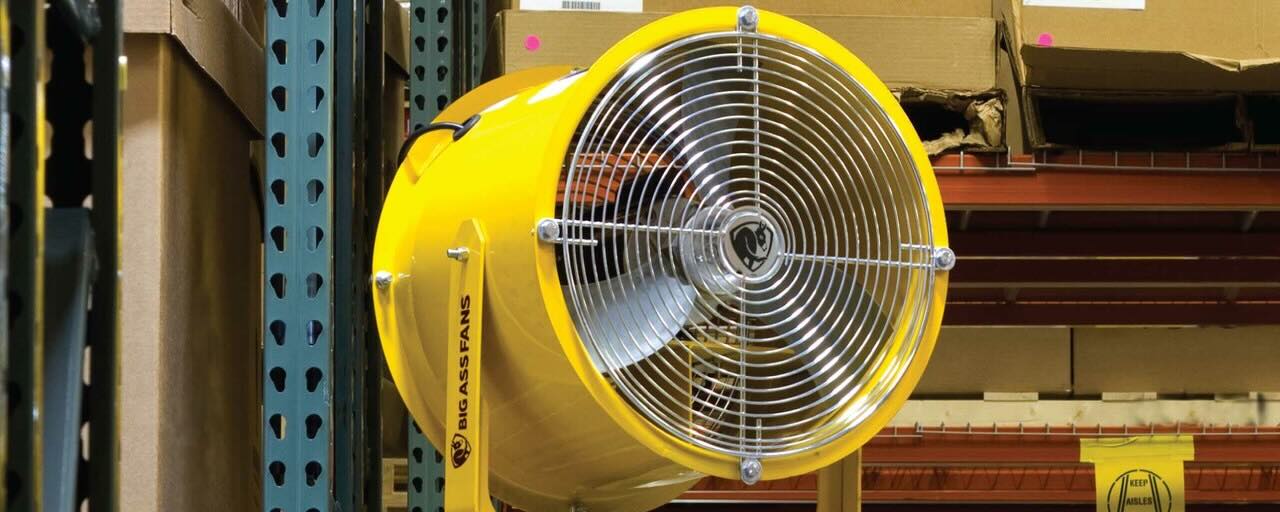 Inside a warehouse, with shelves covered in boxes and wooden pallets, all cooled by a world-renown heavy-duty industrial Big Ass Fan.