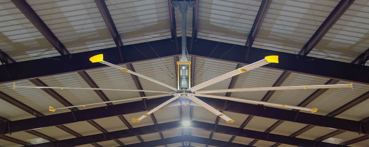 Looking up toward the ceiling inside a large warehouse, with ceiling beams and a large fan by Big Ass Fans.