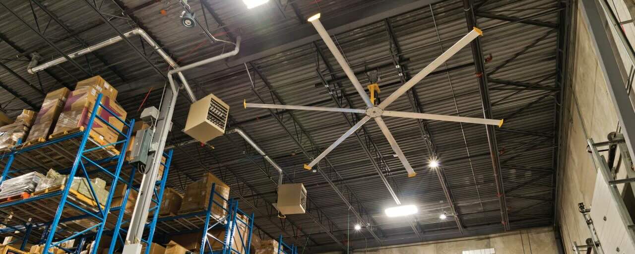 Inside a warehouse facility with many tall shelving units holding heavy boxes and materials. A large HVL fan by Big Ass Fans is positioned to optimize airflow.