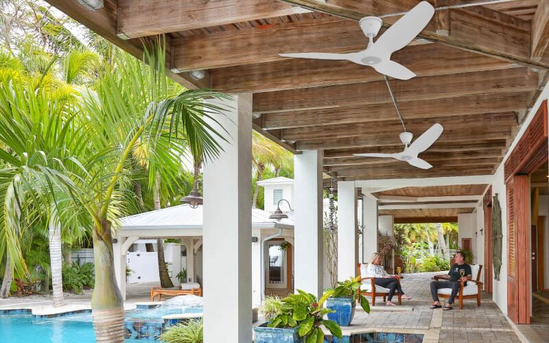 Looking at a tropical resort setting, with a couple relaxing in lounge chairs by the pool under the comfort of Haiku Coastal ceiling fans to relieve the humidity of the tropical environment.