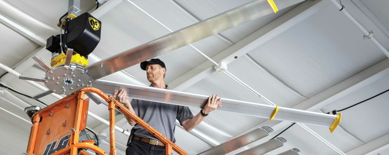 A certified installer for Big Ass Fans on a tall ladder, expertly places a large fan blade into an HVLS ceiling fan.