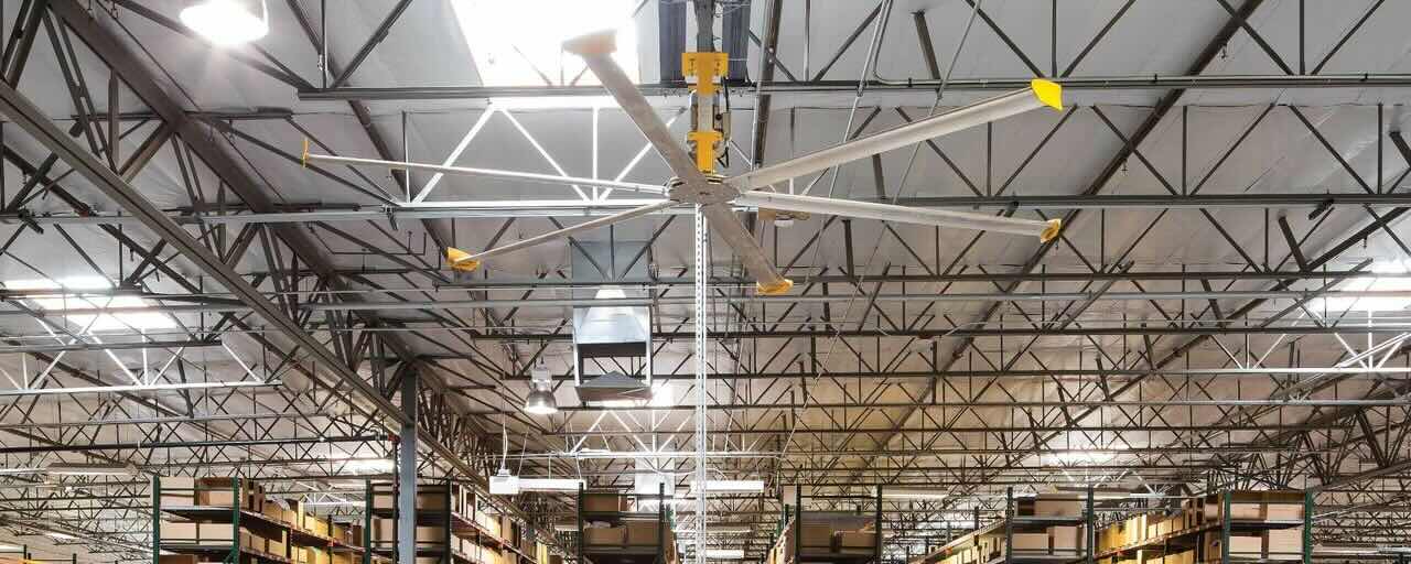 Inside a large warehouse, looking toward the ceiling at an expertly installed HVLS Fan by Big Ass Fans circulating air around large shelves and other obstacles.