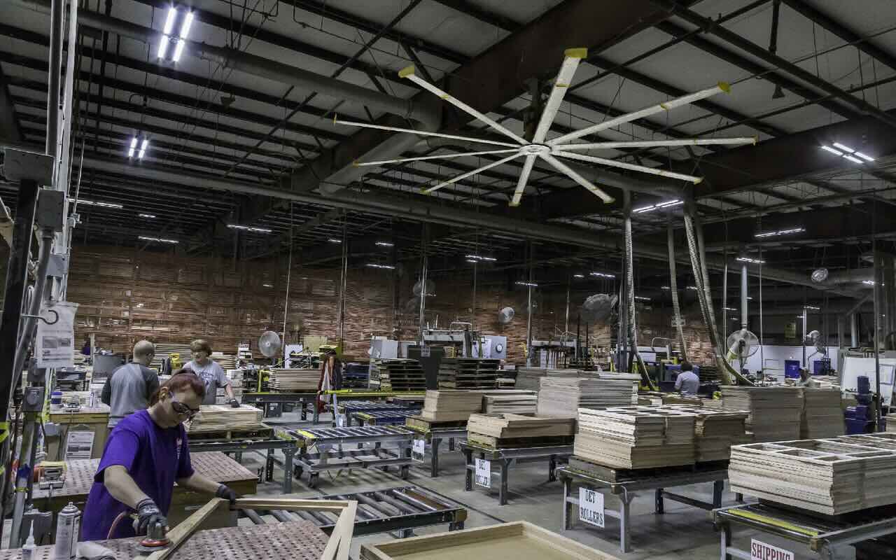 Inside a bustling industrial building full of assembly lines and materials, with employees working on various phases of the assembly process while cooled by the expertly positioned Big Ass Fans on the ceiling and mounted on columns.