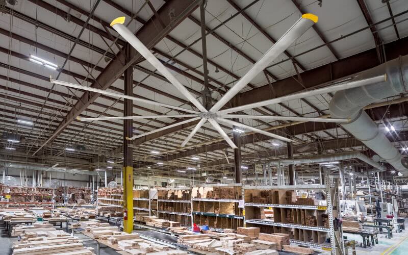 Inside a large warehouse, with many rows of wood and other materials in various stages of processing, a large overhead ceiling fan by Big Ass Fans keeps the air circulating and the environment comfortable for employees.